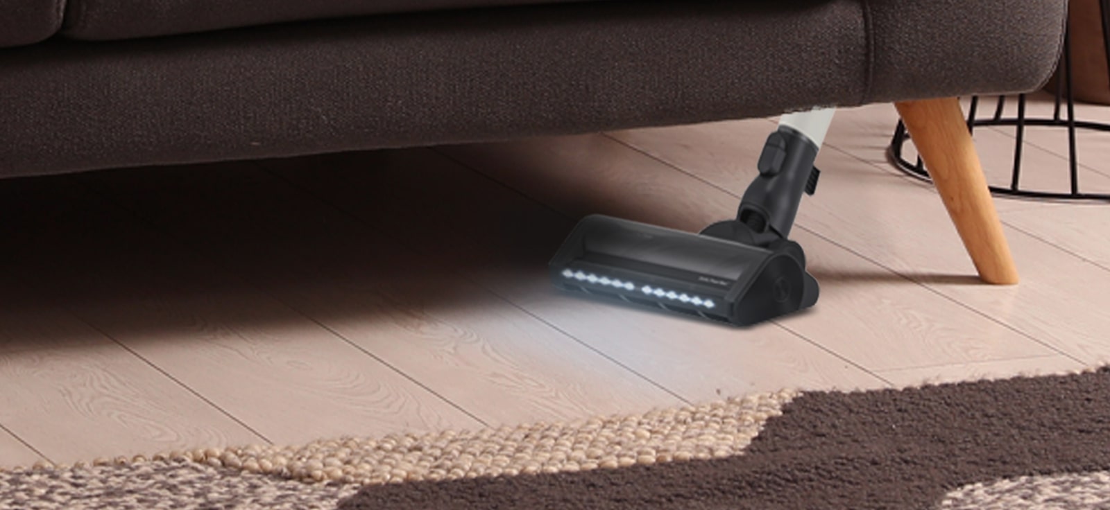 Vacuum cleaner with LED lights cleaning under a brown couch on wooden floor, next to a carpet and a metal basket.